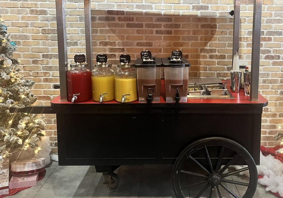 A Bloody Mary cart on a brick wall, jugs of juice and garnishes on a black cart.