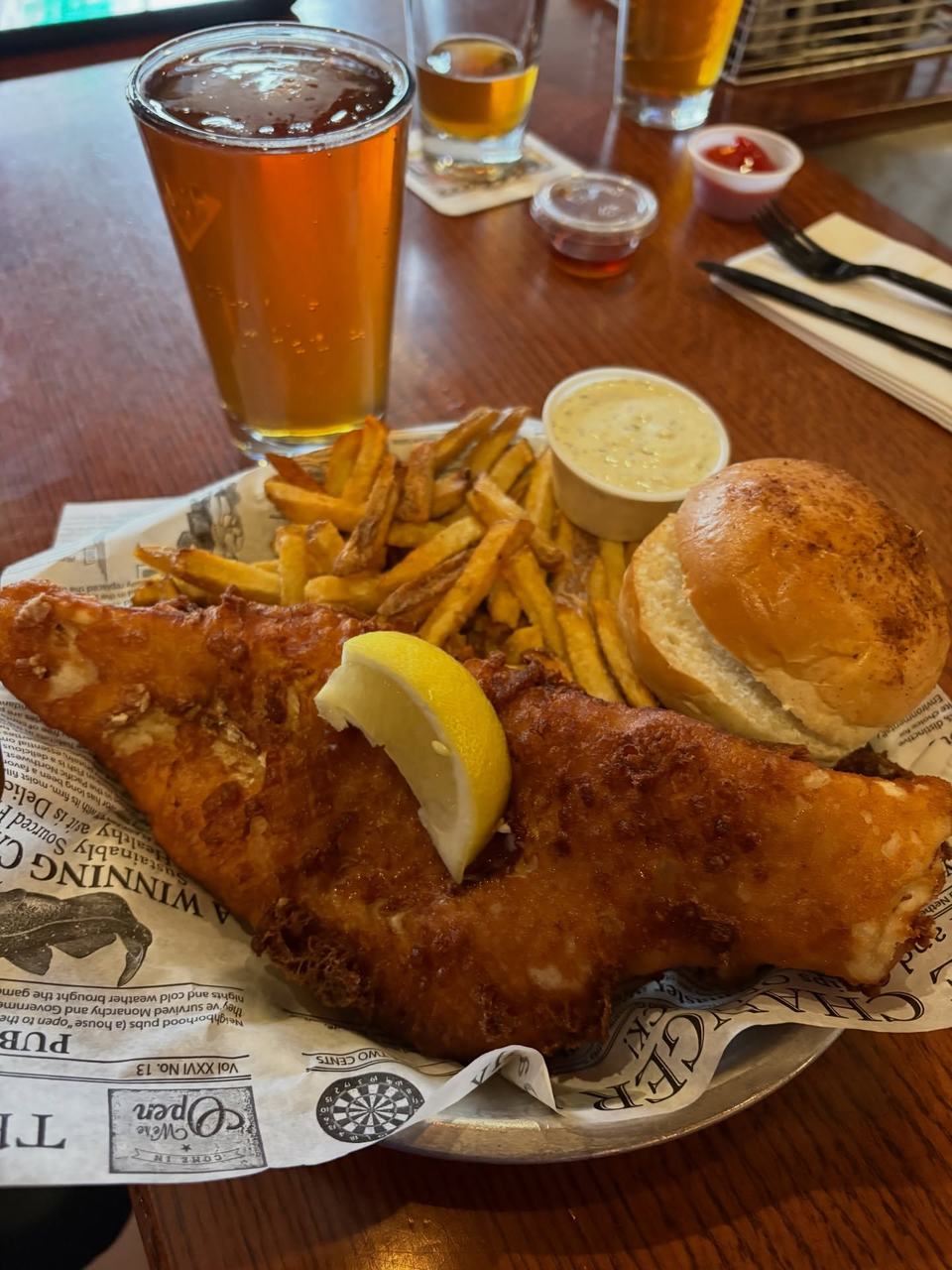 A large beer-battered fish fillet with a lemon wedge, fries, a tartar cup and a roll on newsprint-lined paper, an amber pint behind.