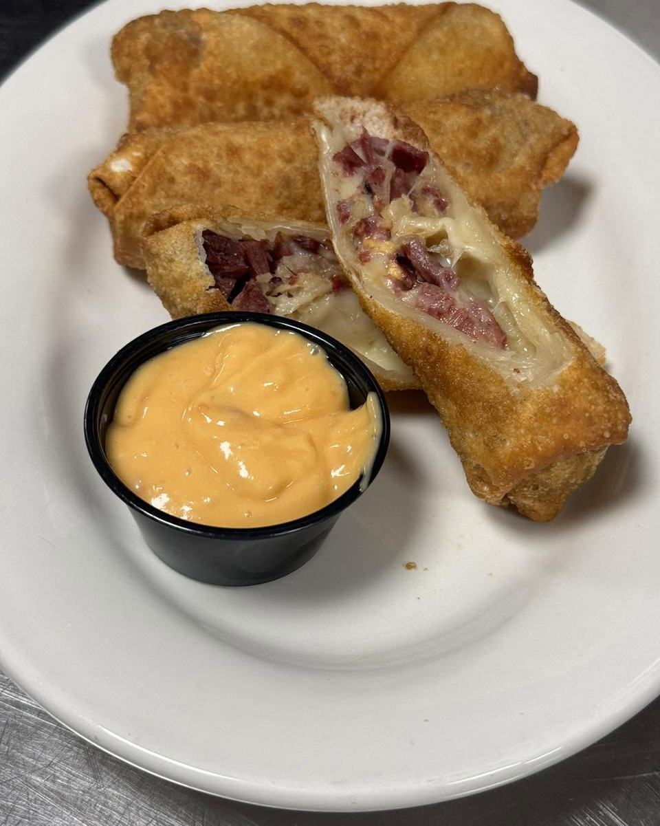 Fried wonton rolls cut open to show corned beef and Swiss, with a cup of orange dipping sauce, on a white plate.
