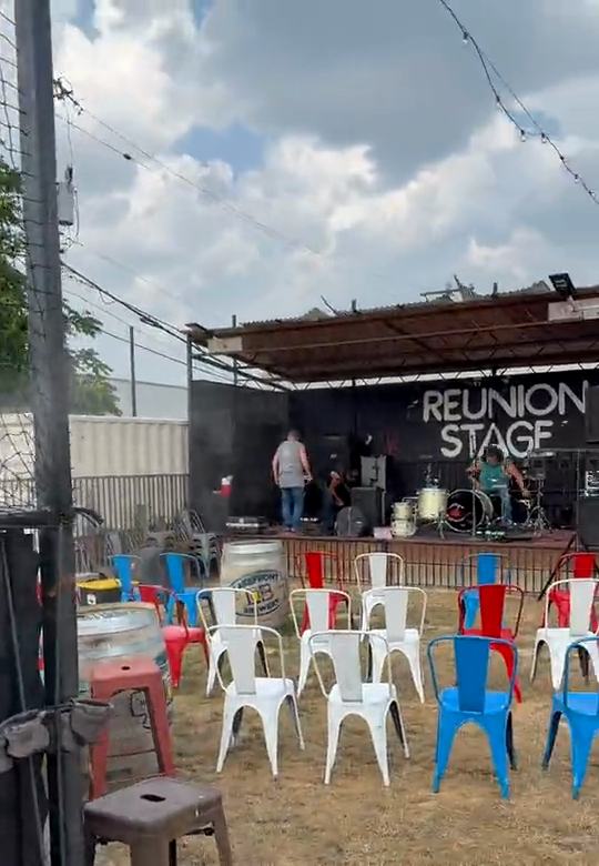 A pan from a misting fence to the daytime lawn and the REUNION STAGE, which dissolves into the same stage at night with a band under pink lights.