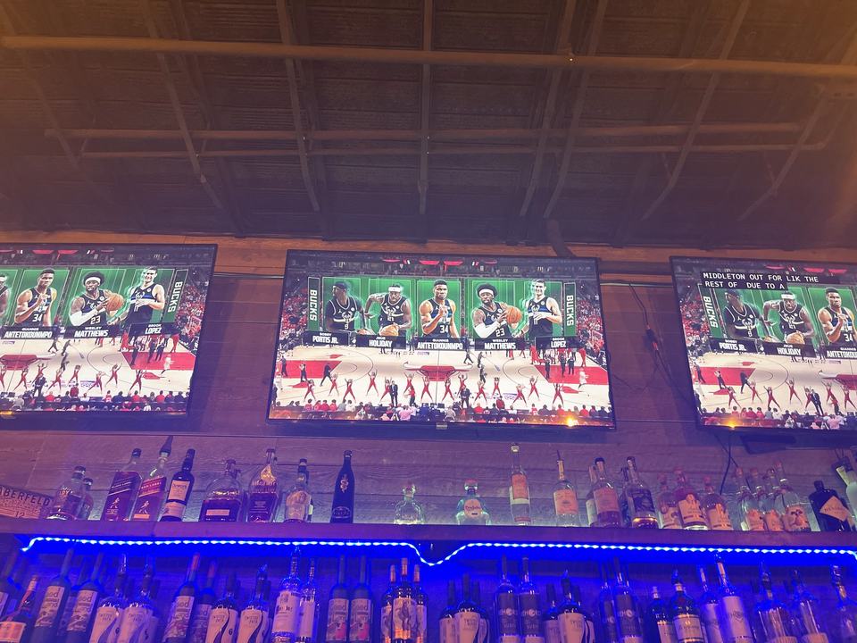 The bar under a wooden ceiling, a row of TVs showing a basketball game above lit liquor shelves.