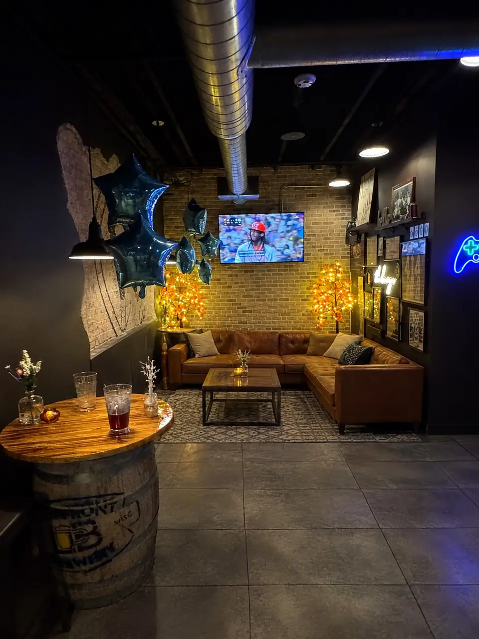 The lounge: a tan leather sectional under lit trees and a TV on the brick wall, a barrel high-top with flowers in front.