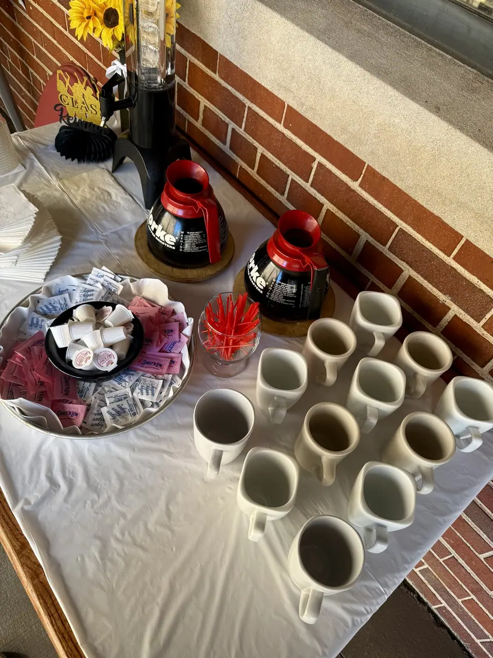 A coffee station on the patio: two pots, rows of white mugs, sugar and creamer.