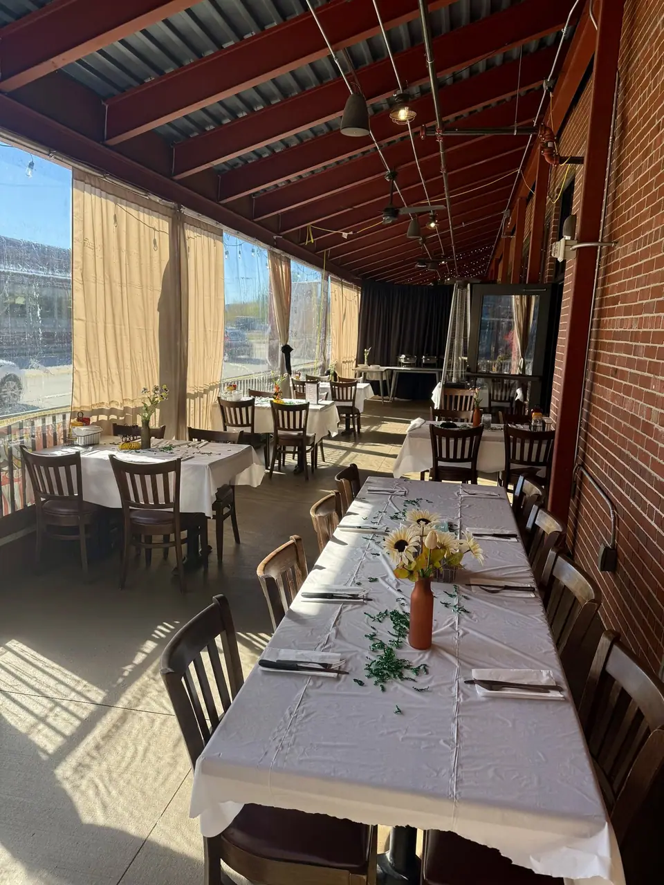 The enclosed patio set for a party: a long table in white linens with flower vases, more tables down the room, curtains along the open side.