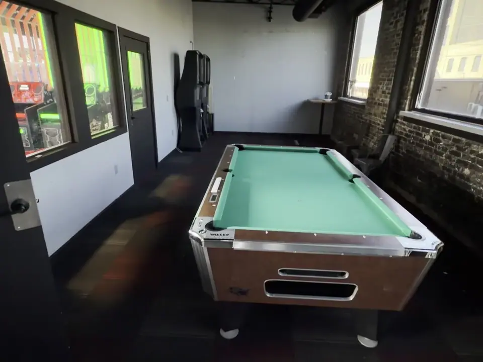 The upstairs party room empty: a pool table, a brick wall with tall windows, and the door to the arcade.