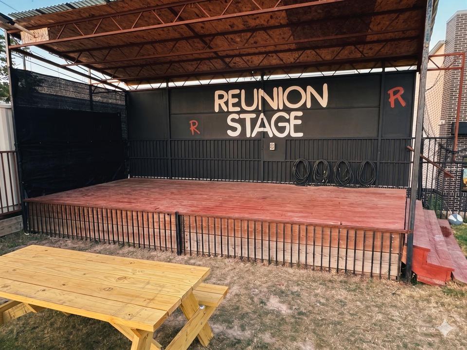 The REUNION STAGE under its roof in daylight, a wooden picnic table in front, no one on stage.